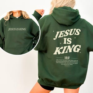 May include: A dark green hoodie with the text "JESUS IS KING" printed on the front and back. The back of the hoodie features the same text in a larger font, along with a crown graphic and a Bible verse.