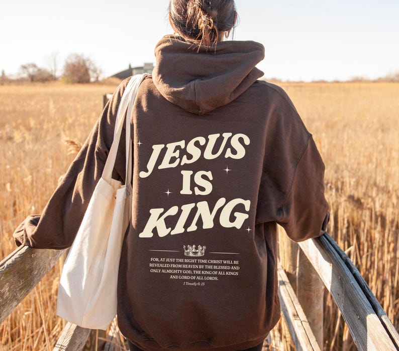 May include: A brown hoodie with the text "JESUS IS KING" in a cream-colored font. Below the text is a quote from 1 Timothy 6:15. The person is wearing a white tote bag and walking on a wooden bridge.