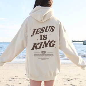 May include: Cream-colored hoodie with the text "JESUS IS KING" in brown lettering on the back. Below the text is a crown graphic and a Bible verse. The hoodie has a hood and long sleeves, suitable for casual wear.