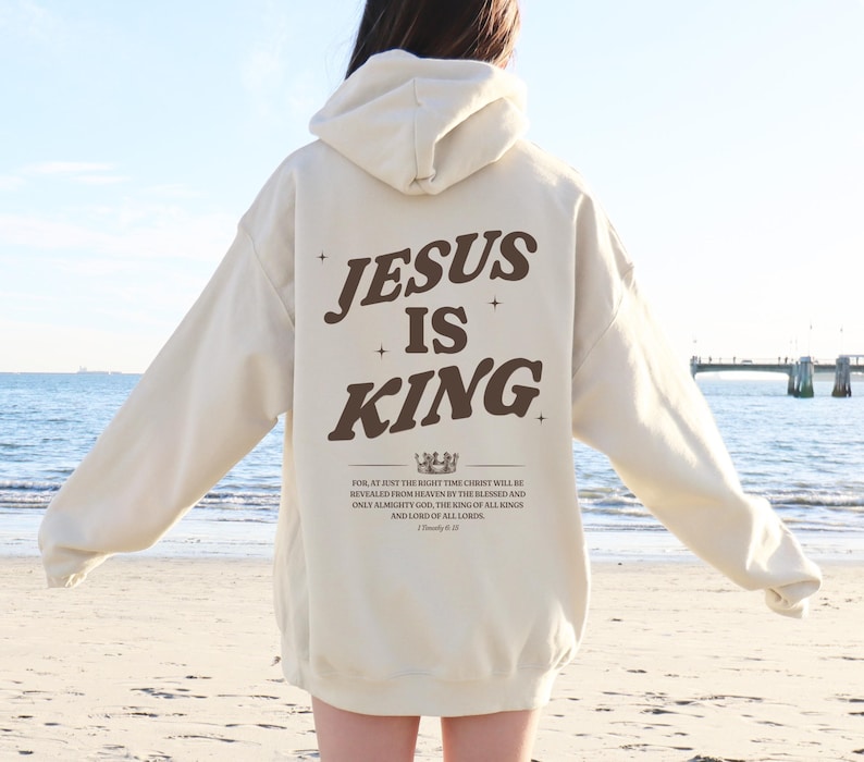 May include: Cream-colored hoodie with the text "JESUS IS KING" in brown lettering on the back. Below the text is a crown graphic and a Bible verse. The hoodie has a hood and long sleeves, suitable for casual wear.