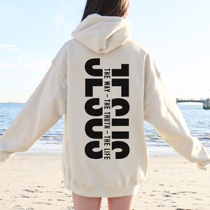 May include: A cream-coloured hoodie featuring the word "JESUS" printed vertically in bold black letters on the back, with the text "THE WAY - THE TRUTH - THE LIFE". The hoodie has a hood and long sleeves.