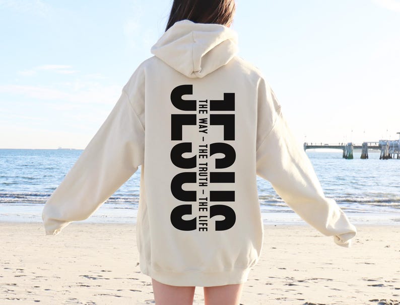 May include: A cream-coloured hoodie featuring the word "JESUS" printed vertically in bold black letters on the back, with the text "THE WAY - THE TRUTH - THE LIFE". The hoodie has a hood and long sleeves.