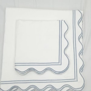 May include: Two white linen napkins with scalloped edges and blue trim. The smaller napkin is placed on top of the larger napkin.