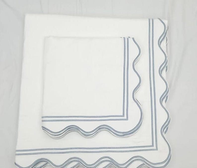 May include: Two white linen napkins with scalloped edges and blue trim. The smaller napkin is placed on top of the larger napkin.