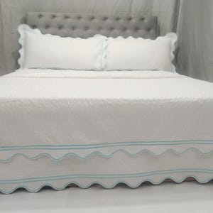Op de afbeelding: Wit bed met een gestoffeerd hoofdeinde en een wit gestikt bedovertrek met een gegolfde rand en lichtblauwe bies. Het bed is opgemaakt met witte kussens met gegolfde randen.