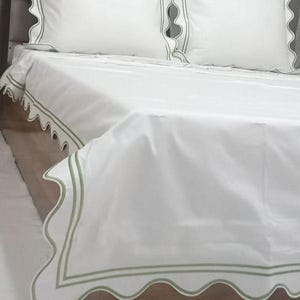 May include: White bed linens with a scalloped edge and green trim. The linens are on a bed with a gray headboard.
