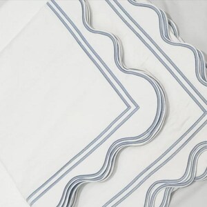 May include: Three white linen napkins with blue scalloped edges and blue stripes. The napkins are folded and stacked on top of each other.