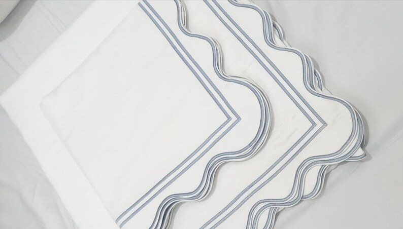 May include: Three white linen napkins with blue scalloped edges and blue stripes. The napkins are folded and stacked on top of each other.