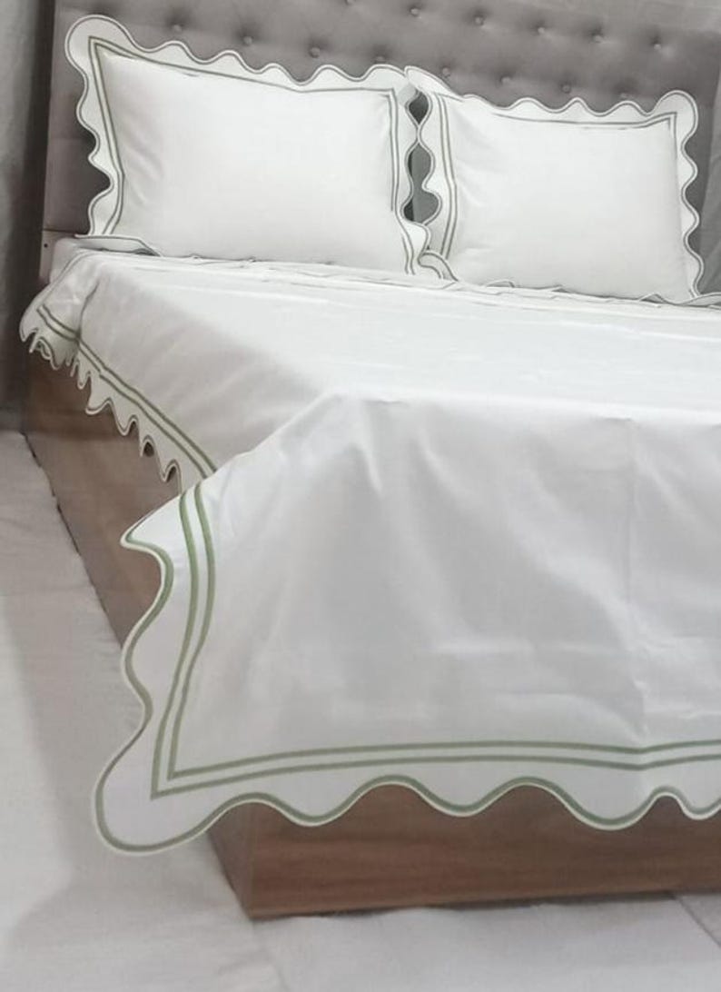 May include: White bed linens with a scalloped edge and green trim. The linens are on a bed with a gray headboard.