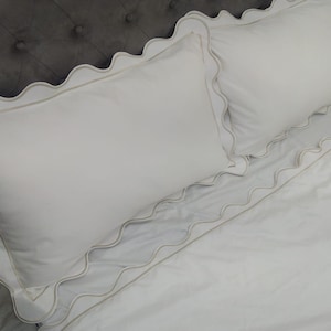 May include: White bedding with a scalloped edge. The bedding is made of a soft, smooth fabric and has a subtle sheen.