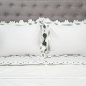 May include: White bed linens with a scalloped edge and a green trim. The linens are on a gray headboard.
