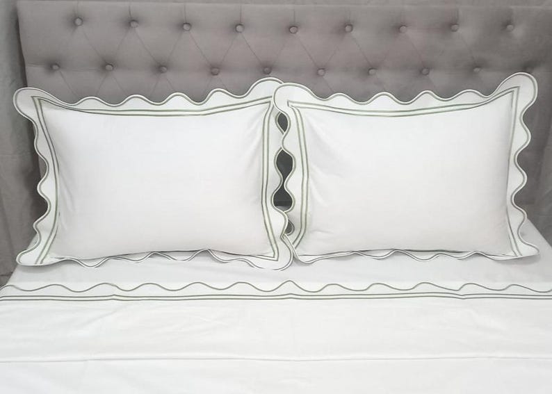May include: White bed linens with a scalloped edge and a green trim. The linens are on a gray headboard.