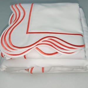 May include: A set of white bed linens with a scalloped edge and a coral colored trim.