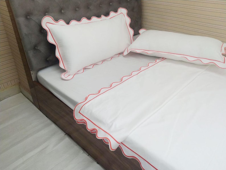 May include: White bed linens with a scalloped edge and a red trim. The linens are on a bed with a brown headboard.
