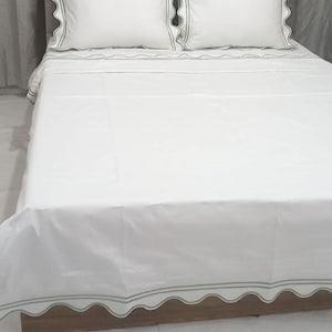May include: White bed linens with a scalloped edge and a green trim. The linens are on a bed with a gray headboard.