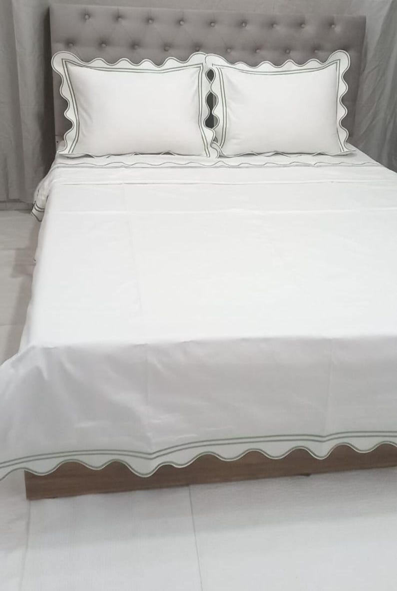 May include: White bed linens with a scalloped edge and a green trim. The linens are on a bed with a gray headboard.