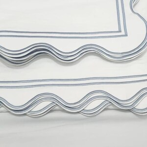 May include: White bedding with a scalloped edge and blue stripes. The bedding is folded in layers, showing the detail of the scalloped edge.