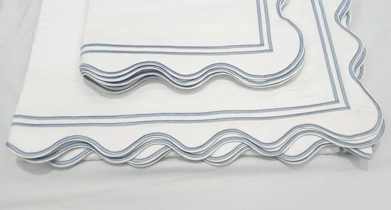 May include: White bedding with a scalloped edge and blue stripes. The bedding is folded in layers, showing the detail of the scalloped edge.