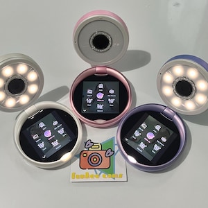 May include: A collection of small, round, selfie-style cameras in various colors, including pink, white, and purple. Each camera has a screen displaying icons and settings, and a ring light. The cameras are arranged around a cartoon camera logo with the text "Funkee cams".
