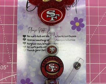 May include: A San Francisco 49ers badge reel featuring a red glittery Mickey Mouse-shaped acrylic topper with a gold glitter bow. The lanyard includes beads with the 49ers logo, a football graphic, and red and white accents on a clear clip.