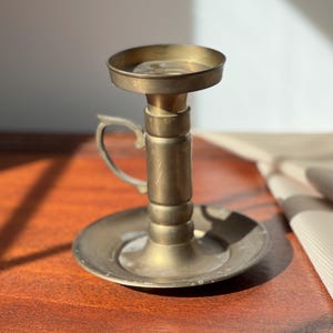 May include: A vintage brass candlestick holder with a round base and a small handle. The candle holder has a circular top to catch wax. The brass has a warm, aged patina, suggesting it is an antique.