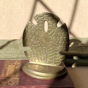 May include: A brass bookend shaped like a sand dollar with a detailed design. The bookend is sitting on top of a red book with gold lettering that reads "Scott's Marmion".