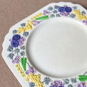 May include: A white ceramic plate with a floral design. The plate has a scalloped edge and is decorated with purple, yellow, and green flowers and leaves. The flowers are arranged in a repeating pattern around the rim of the plate.