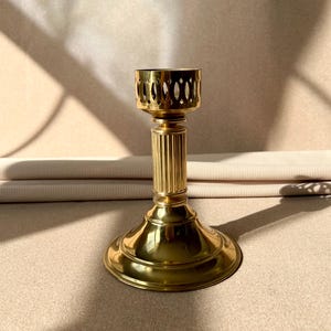 May include: A gold metal candle holder with a fluted column and a decorative top. The holder is designed to hold a single candle.