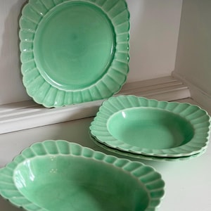 May include: A collection of mint green ceramic plates and bowls with scalloped edges. The set includes a large round plate, a square plate, and an oval bowl. The dishes are displayed on a white shelf.
