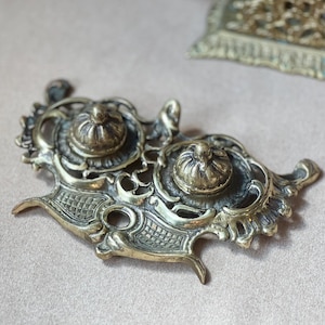 May include: Ornate antique brass inkwell with two ink pot holders and decorative filigree. The inkwell has a detailed, aged appearance, with a textured surface and intricate floral designs. The overall shape is asymmetrical with curved edges and small feet.