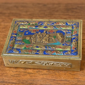 May include: A rectangular brass box featuring a hinged lid with colorful enamel inlays of blue, green, and red. The lid displays a central relief scene of figures in an outdoor setting, surrounded by a decorative border of floral and leaf motifs.