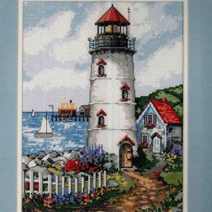 Könnte beinhalten: Eine Kreuzstich-Stickerei, die einen weißen Leuchtturm mit rotem Dach, ein kleines Haus mit rotem Dach und einen weißen Lattenzaun zeigt. Der Leuchtturm steht an einem felsigen Ufer mit blauem Wasser und einem Segelboot in der Ferne. Der Himmel ist blau mit weißen Wolken. Der Text "LADY DIAMONDS EMPORIUM" befindet sich oben im Bild.