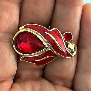 May include: A gold-tone brooch featuring a large, teardrop-shaped red faceted crystal centerpiece. The metal frame is accented with smooth, vibrant red enamel sections and a small, circular red stone detail on the right side of the design.