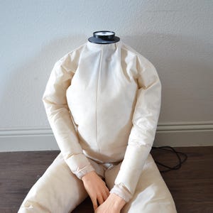 Puede incluir: Maniquí sin cabeza en posición sentada, con un traje acolchado color crema. El maniquí tiene manos de aspecto realista y una pieza negra circular en el cuello. El traje está sobre un suelo de madera oscura.