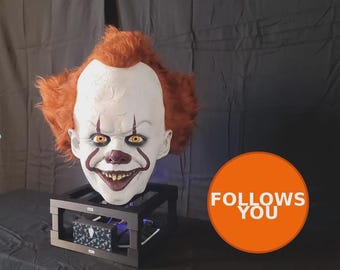 May include: A detailed, realistic clown head with orange hair, white face paint, and a sinister grin. The clown has yellow eyes and red accents around the mouth and eyes. The head is mounted on a black stand, with the text "FOLLOWS YOU" in an orange circle.