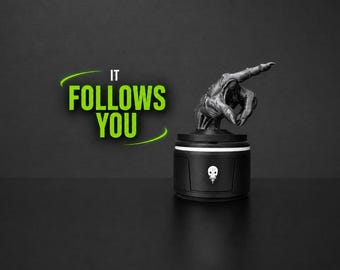 May include: A black cylindrical grinder featuring a sculpted dark gray hand reaching upward from the lid. The side of the grinder displays a small white skull icon. The text IT FOLLOWS YOU is printed in bright green letters to the left of the object.