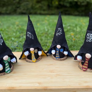 Peut inclure: Quatre gnomes faits à la main avec des chapeaux pointus noirs, chacun représentant une maison différente d'une série fantastique. Les chapeaux ont des symboles brodés blancs. Les gnomes ont des cravates colorées et sont exposés sur des rondins de bois.