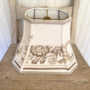 May include: An off-white, octagonal lampshade featuring a beige embroidered floral pattern around the lower section. The shade has a structured fabric exterior with a brass-toned metal spider fitter at the top center for attachment to a lamp base.