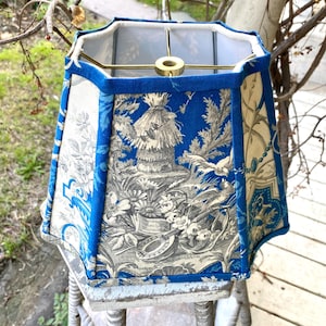 May include: An octagonal lampshade with a blue and cream toile pattern. The shade features a vibrant blue trim and a gold-colored metal frame. The toile design depicts scenes of nature and figures.