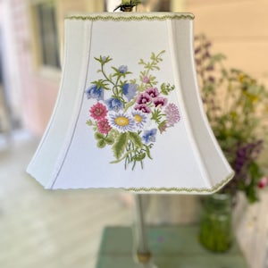 May include: A white fabric lampshade featuring a central embroidered floral bouquet with blue, pink, and white flowers and green leaves. The shade has a flared rectangular shape with a decorative green leaf-patterned trim along the top and bottom edges.