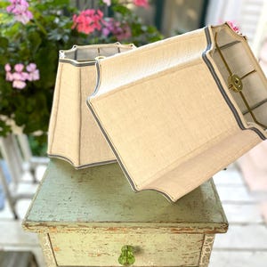 May include: Two off-white fabric lamp shades with a bell-shaped silhouette and scalloped edges. Each shade features a thin, dark blue trim along the borders and a brass-toned metal spider fitter inside the top opening for attachment to a lamp base.