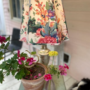 May include: A table lamp with a pleated, cream-colored fabric shade featuring a vibrant, multi-colored floral pattern of pink, orange, and green botanical illustrations. The lamp has a metallic, silver-toned base and a central stem.