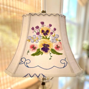 May include: A cream-colored fabric lampshade featuring a central embroidered floral design of yellow, purple, and pink pansies with green leaves. The shade has a scalloped top edge with blue stitching and blue decorative scrollwork at the base.