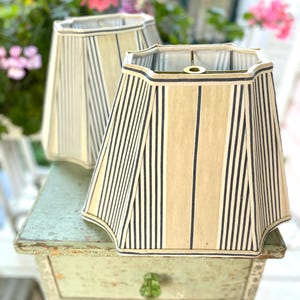 May include: Two bell-shaped lamp shades featuring a vertical striped pattern in navy blue and beige. The shades have a scalloped bottom edge and a structured frame with a central metal spider fitter at the top for attachment to a lamp base.