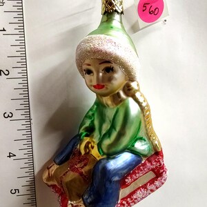 May include: A glass Christmas ornament shaped like a child in a green hat and sweater sitting on a red sled, holding a gold gift. A ruler next to the ornament shows it is approximately 5.5 inches tall. A pink tag with the number 560 is attached.