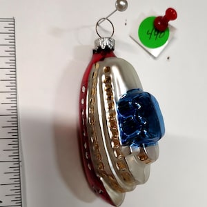 May include: A vintage Christmas ornament shaped like a silver and red airplane with a blue gem-like accent. The ornament has a silver metal loop for hanging and is attached to a white surface.