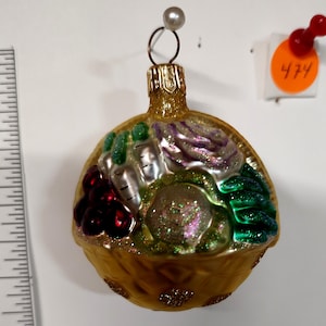 May include: A glass Christmas ornament shaped like a basket filled with colorful vegetables. The ornament features a gold-colored basket with a variety of vegetables, including red grapes, green lettuce, and purple cabbage. The ornament has a gold cap and a pearl on top.