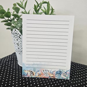 May include: A lined notepad with a seashell design at the bottom. The notepad is white with black lines and a decorative border featuring various seashells in shades of blue, pink, and beige. A small white bucket with green plants is in the background.