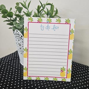 May include: A white notepad with a pink border and the words "to do list" in blue script. The notepad is decorated with yellow pineapples and green leaves. The notepad has lined pages for writing.