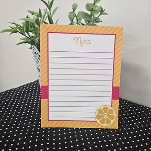 May include: A lined notepad with a citrus theme. The notepad has a white interior with black lines and the word "Notes" in cursive. The border is orange with pink accents and a lemon slice graphic.